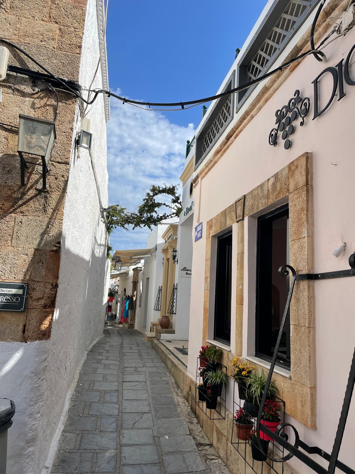Auf dem Foto ist eine Gasse in Lindos zu sehen. Auf der rechten Seite sind zwei Fenster zu sehen. Diese sind dunklebraun die Hauswand ist weiß. Dahinter lassen sich gleichfarbige Türen erahnen.