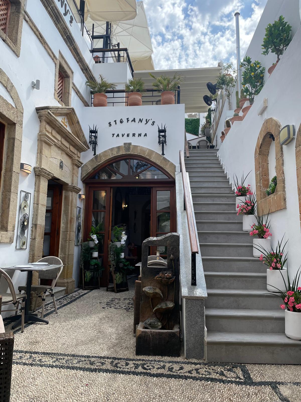 Auf dem Foto ist auf der linken Seite der Eingang einer Taverne zu sehen und auf der rechten Seite eine Treppe, die hinauf auf einen Balkon über der Taverne führt.