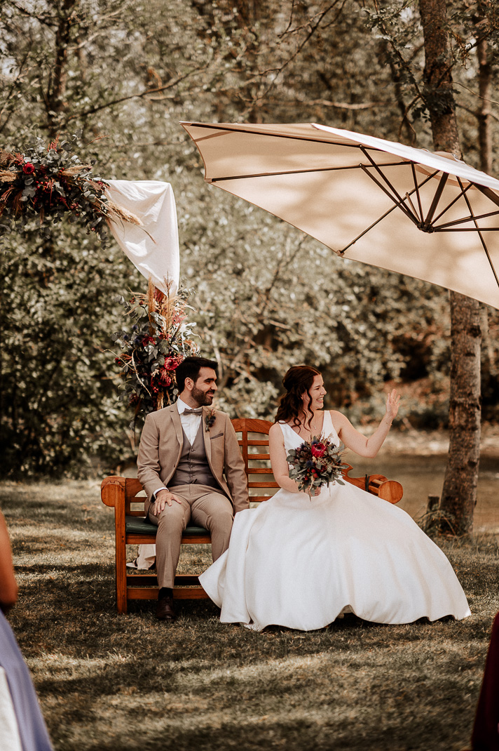 Ein Brautpaar sitzt auf einer Bank bei seiner Trauung und winkt.