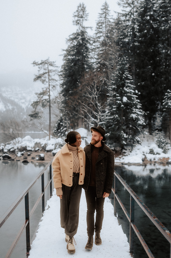 Ein Liebespaar steht auf einer Brücke am winterlichen Eibsee. Pärchenfotos für Einladungskarten zur Hochzeit. 