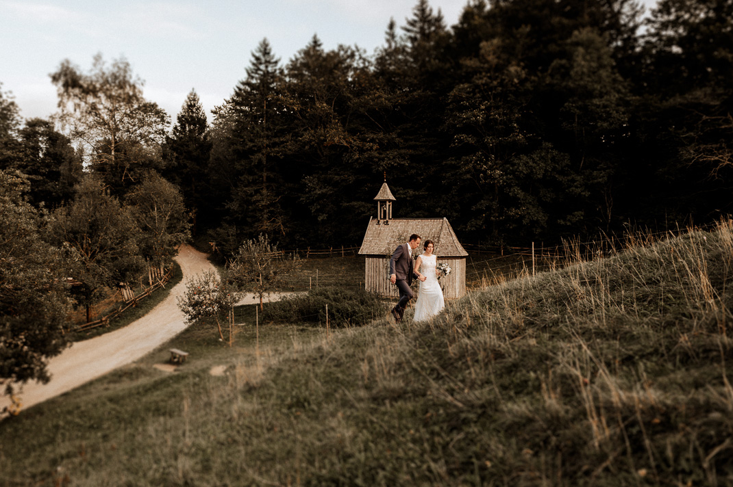 Brautpaarfotos von Hochzeitsfotograf Andreas Reiter. Das Brautpaar geht einen Berg hoch, im Hintergrund ist eine Kapelle zu sehen. Brautpaarfotos beim Wasmeier Freilichtmuseum am Schliersee. 