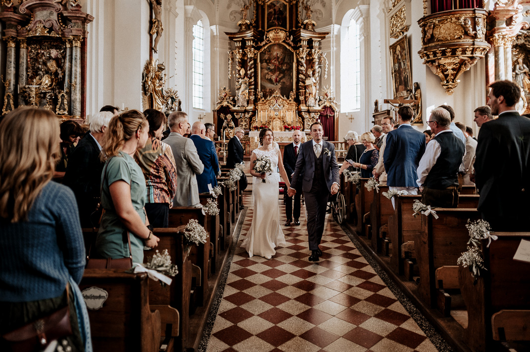Hochzeitsfotograf in der St. Sixtus Kirche am Schliersee. Das Brautpaar zieht Hand in Hand aus der Kirche aus. Reportagefoto von der Trauung.