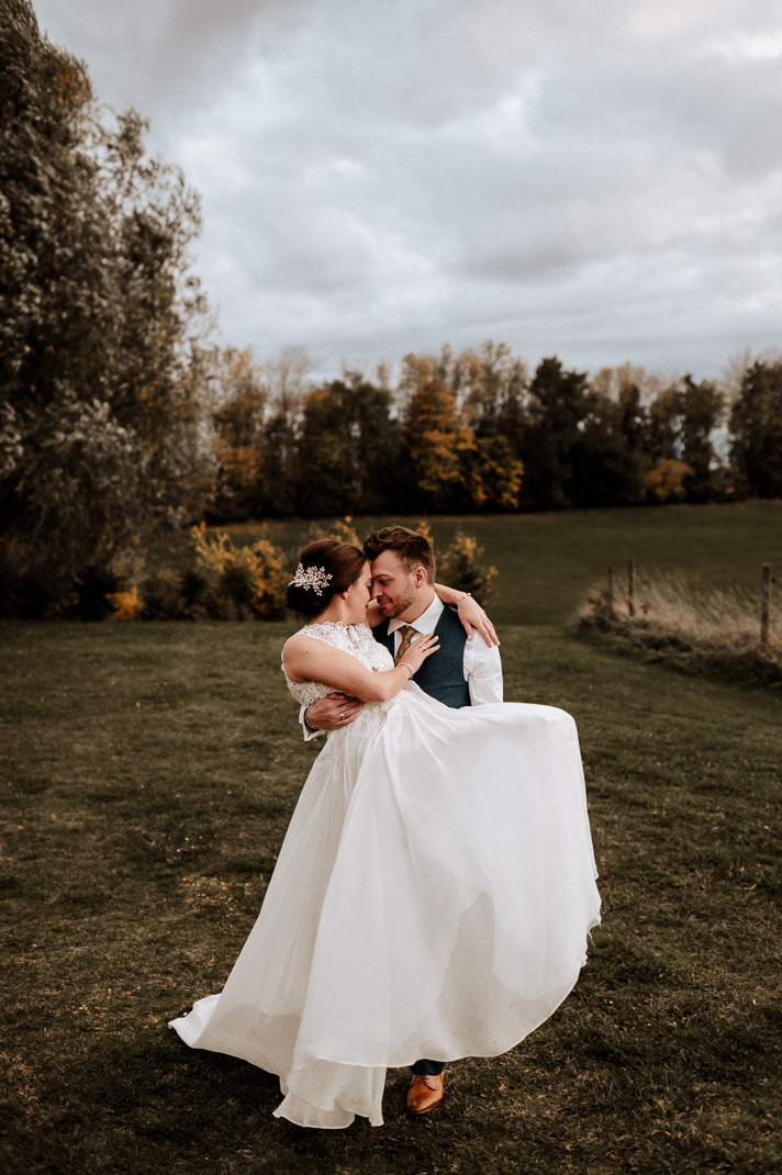 Der Bräutigam hebt die strahlende Braut hoch vor einem herbstlichen Hintergrund.