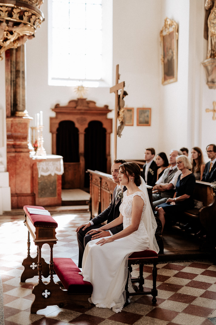 Hochzeitsfotograf in der St. Sixtus Kirche am Schliersee. Reportagefoto von der Trauung. Das Brautpaar sitzt vor dem Altar.