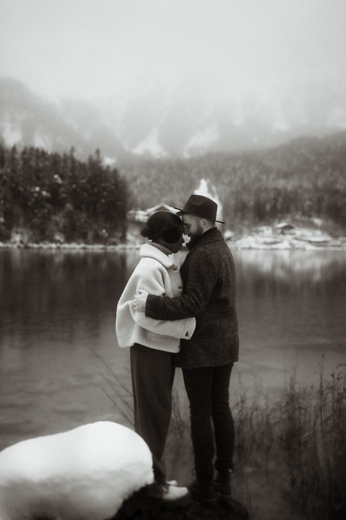 Beim Paarshooting steht ein Paar auf einem Stein im Eibsee, sie umarmen sich fest. Man kann die verschneite Zugspitze im Hintergrund sehen.