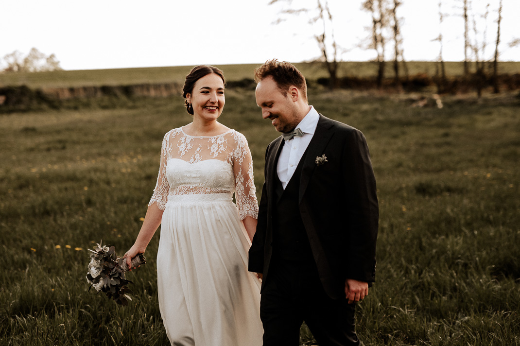 Paarfotos auf einer Wiese bei Zolling. Die Braut sieht zu ihrem Bräutigam, beide gehen über eine Wiese. Hochzeitsfotograf ist Andreas Reiter.