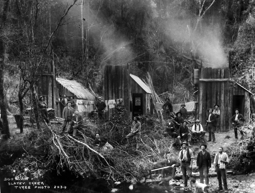 Men (probably tree fellers) in the bush at Dogtown, Slaty Creek, Taitapu. They stand by a river bed next to three small dwellings.  (c.1890s Tyree Studio - Photo of photo in Mangakarau Community Centre). 