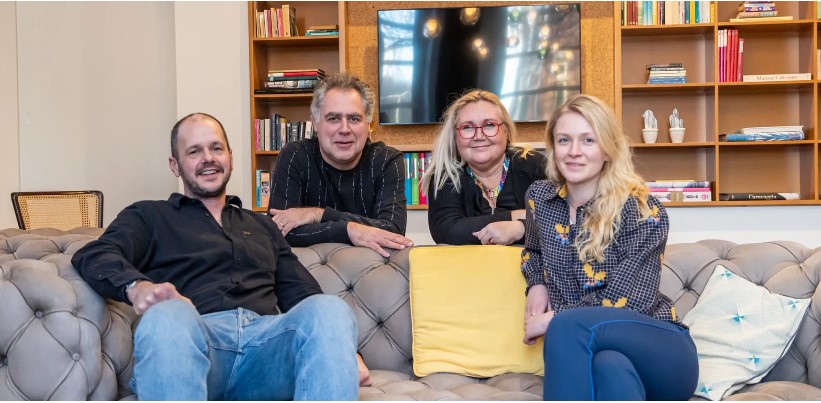 Ein Mann und eine Frau mit langen blonden Haaren sitzen auf einem Sofa und lächeln in die Kamera. Ein weiterer Mann und eine weitere blonde Frau befinden sich hinter dem Sofa, stützen sich auf der Sofalehne ab und lächeln ebenfalls in die Kamera. 