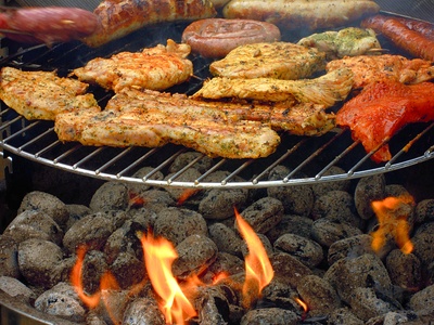 Fleischstücke und Würstchen liegen auf einem Grillrost, das über einem Feuer hängt.