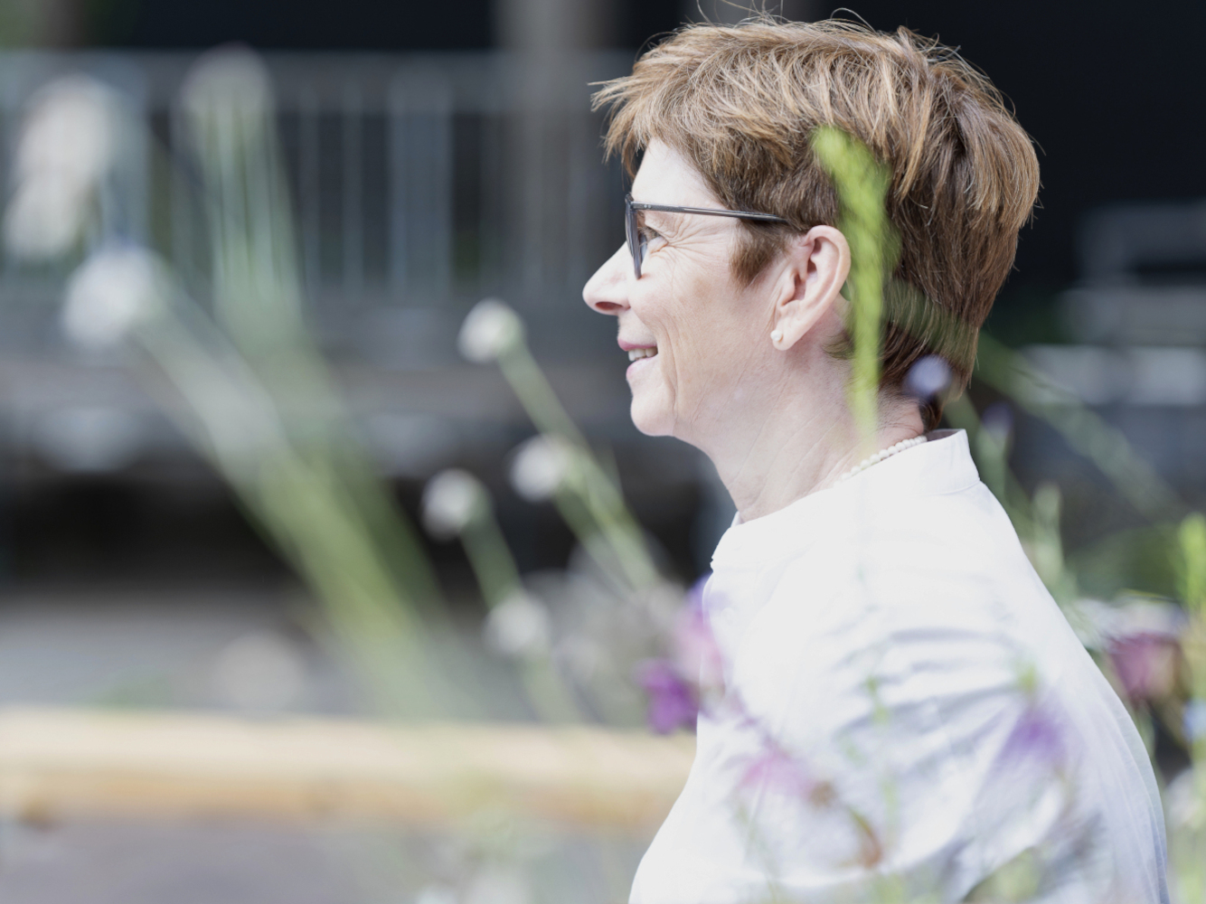Webseite-Porträt einer älteren Frau mit einem weißen Kleid sitzt draußen in einem Hof und schaut nach links. Sie ist im Profil abgebildet. Im Vordergrund sind unscharf rosa Blumen zu sehen. 