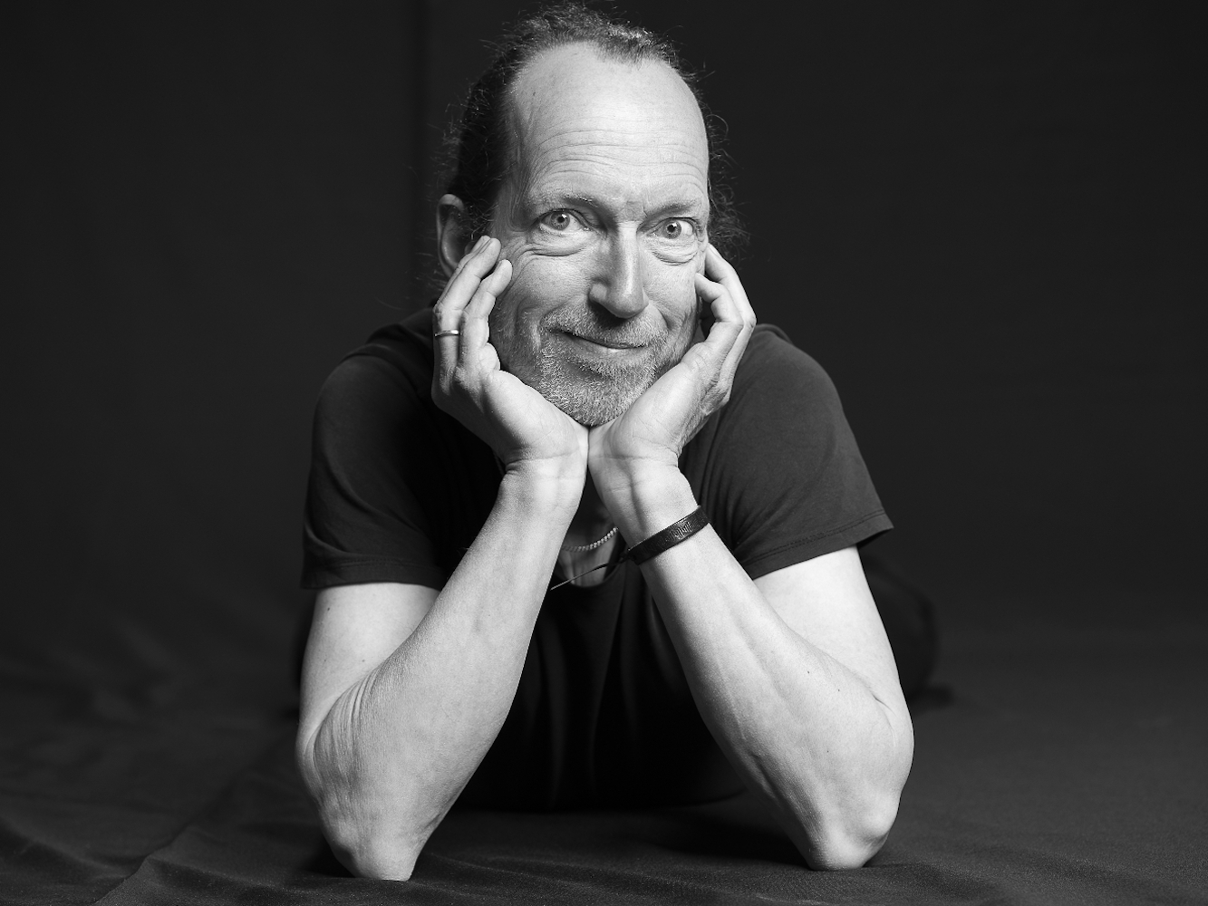 Webseite-Porträt des Pianisten Eckhard Wiemann. Er hat einen 3-Tage-Bart und trägt ein schwarzes T-Shirt. Er liegt auf einem dunklen Untergrund. Er stützt das Kinn in die Ellenbogen und lächelt verschmitzt.