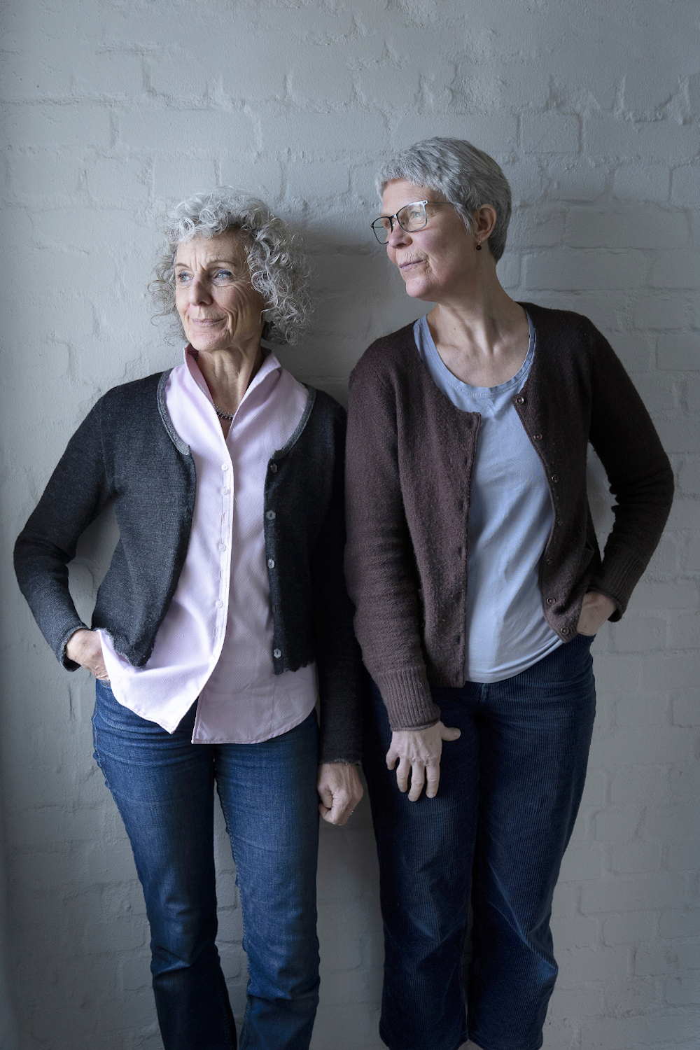 Ina Hattebier und Ute Arndt haben in Hamburg das Netzwerk Trauerkultur gegründet. Die beiden älterern grauhaarigen Frauen stehen im Treppenhaus vor einer weißen Ziegelwand und sehen beide nach links aus dem Fenster.