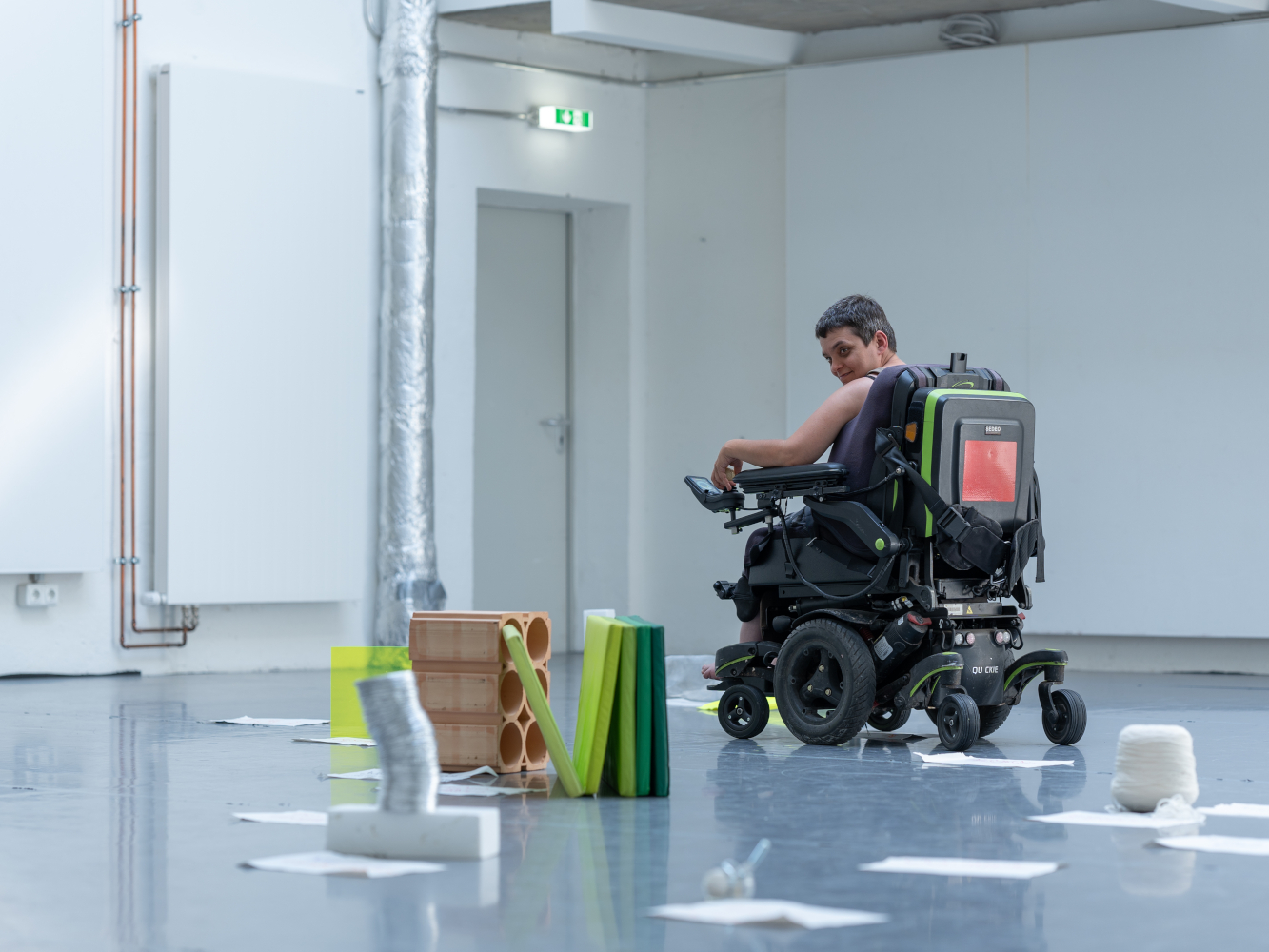 Porträt der Schauspielerin Canan Salman bei einer Theaterprobe. Sie sitzt in einem E-Rolli und fährt zwischen verschiedenen Requisiten und Papierblättern, die auf dem Boden liegen. Sie blickt schräg nach unten, der Blick konzentriert und leicht amüsiert.