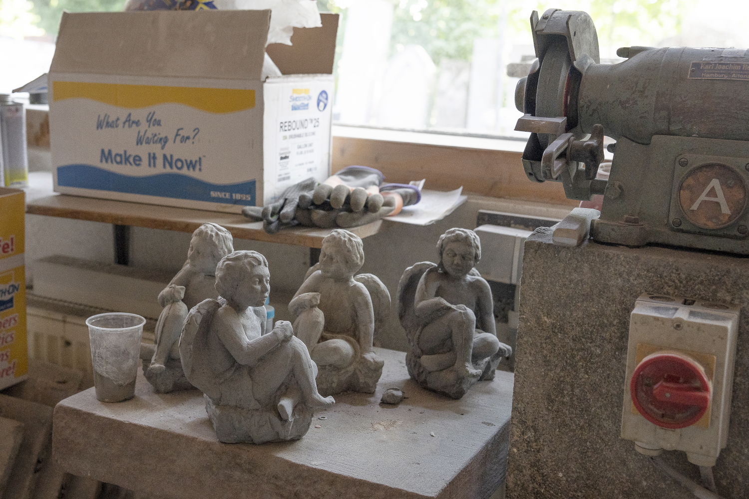 Ein Blick in die Werkstatt des Steinmetzbetriebes Carl Schütt und Sohn. Neben einem Schleifstein und Pappkisten stehen auf einer dicken Steinplatte viele kleine Engelfiguren. Es sieht aus, als ob die Engel sich unterhalten..