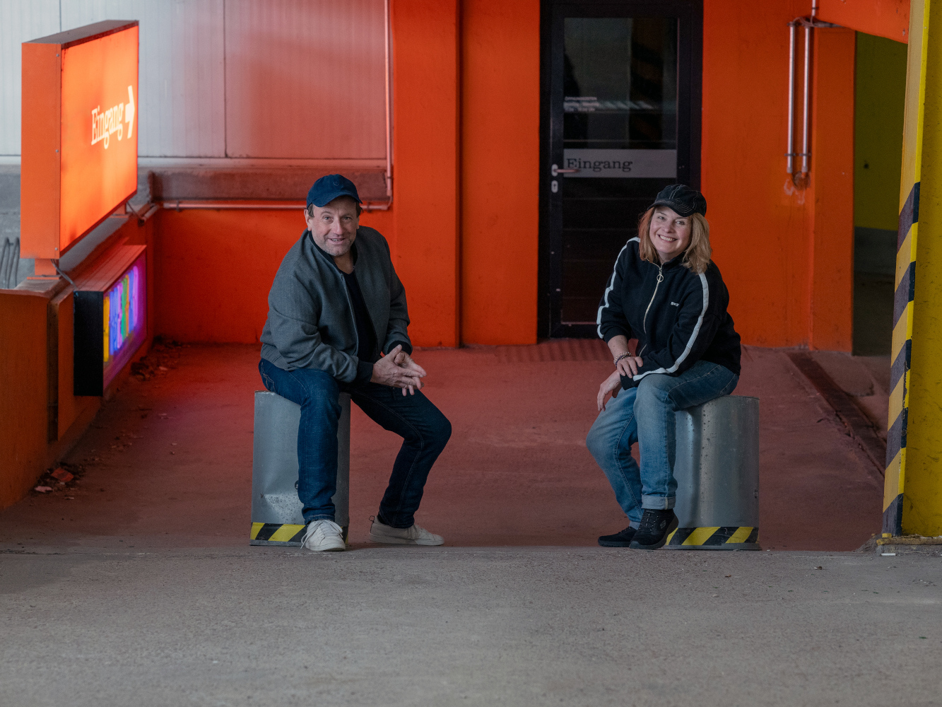 Porträt von Peter G.Dirmeier und Silke Roca (Theater Echtzeit). Die beiden sitzten in auf zwei umgedrehten Blecheimern in einem Parkhaus. Im Hintergrund ist eine orange Wand. Neben den beiden ist ein orangenes Leuchtschild mit der Aufschrift Eingang.   