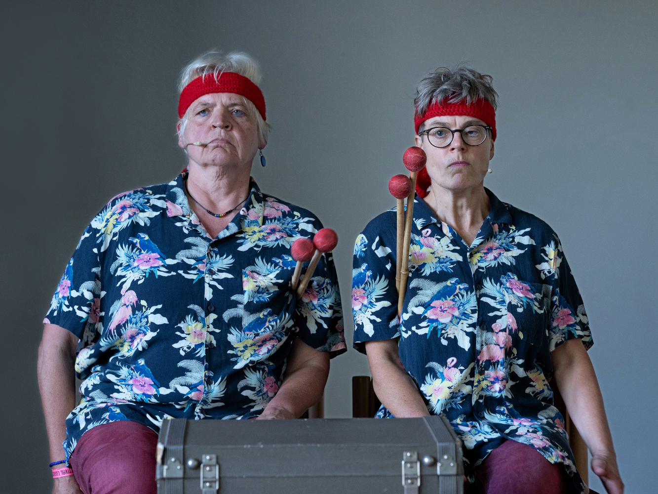 Porträt von Annette Kayser und Keo Hundius vom Rhythmusikabarett A-Quadrat. Sie tragen wild gemusterte Hemden und rote Stirnband. Sie sitzen mit verkniffenen Gesichtern hinter einem Koffer und haben je 2 Klöppel mit roten Enden unter den Arm geklemmt.