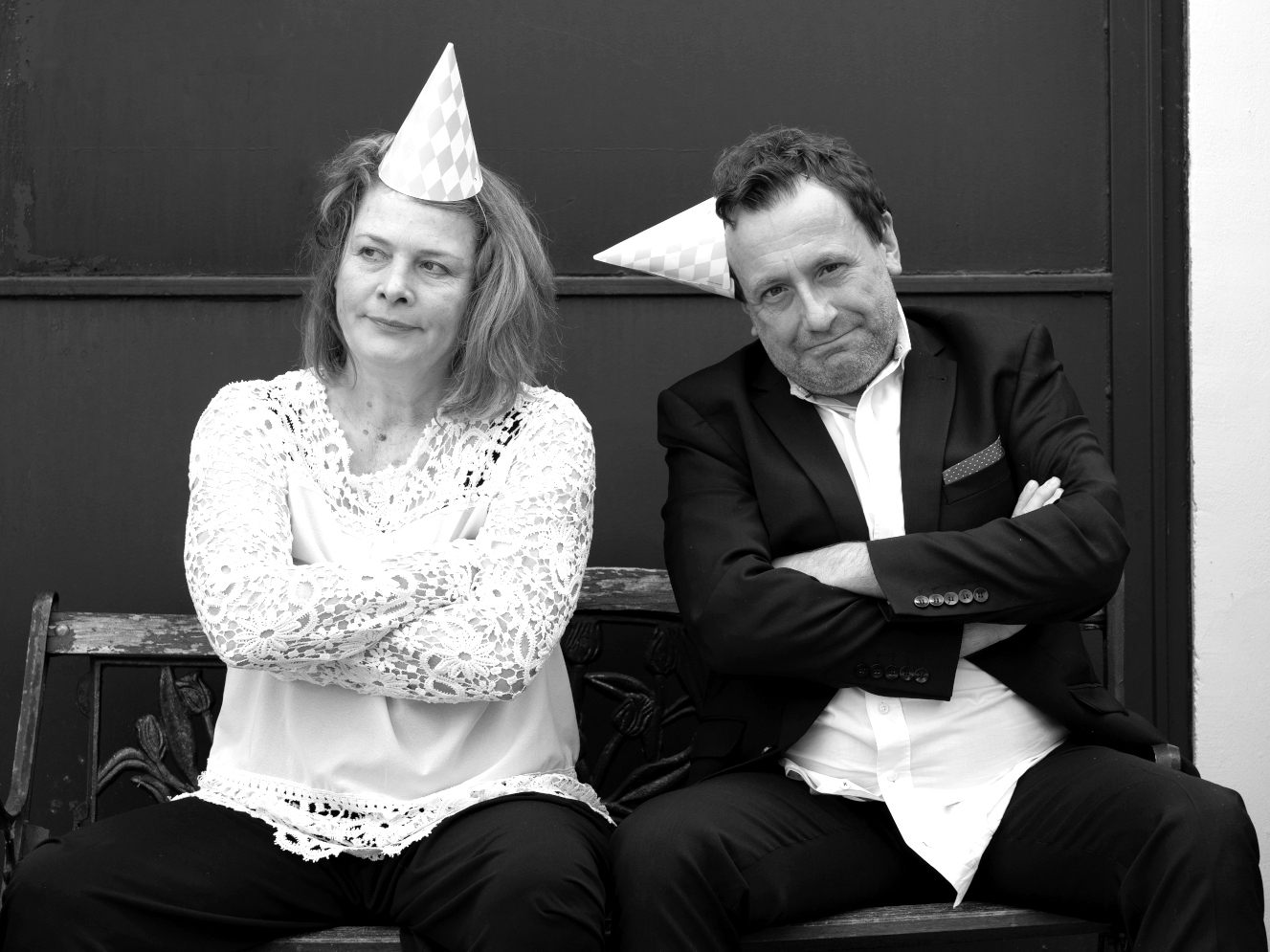 Schwarzweiss-Porträt von Peter G.Dirmeier und Silke Roca (Theater Echtzeit). Die beiden auf einer Bank, sie wirken genervt voneinander. Sie tragen Papphütchen. Bei ihm ist das Hemd aus der Hose gerutscht und das Hütchen sitzt wie ein Horn auf dem Ohr.