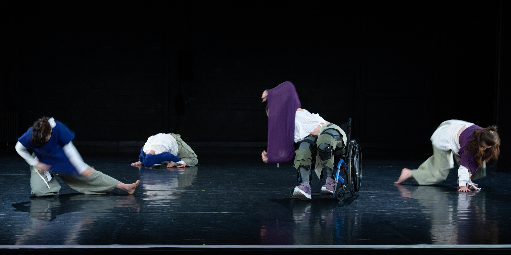 Vier Tanzende auf einer dunklen Bühne winden sich wütend am Boden oder im Sitzen aus ihren blauen und lila Oberteilen. Eine steckt mit dem Kopf im blauen Stoff fest, eine andere wirkt, als kippe sie bei ihrer wilden Bewegung gleich aus dem Rollstuhl.