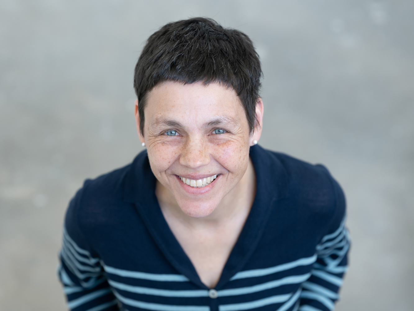 Porträt der Trauerrednerin Annette Rosenfeld. Sie hat kurze dunkle Haar, trägt ein blau gestreiftes Oberteil. Sie sieht nach oben und strahlt über das ganze Gesicht. 