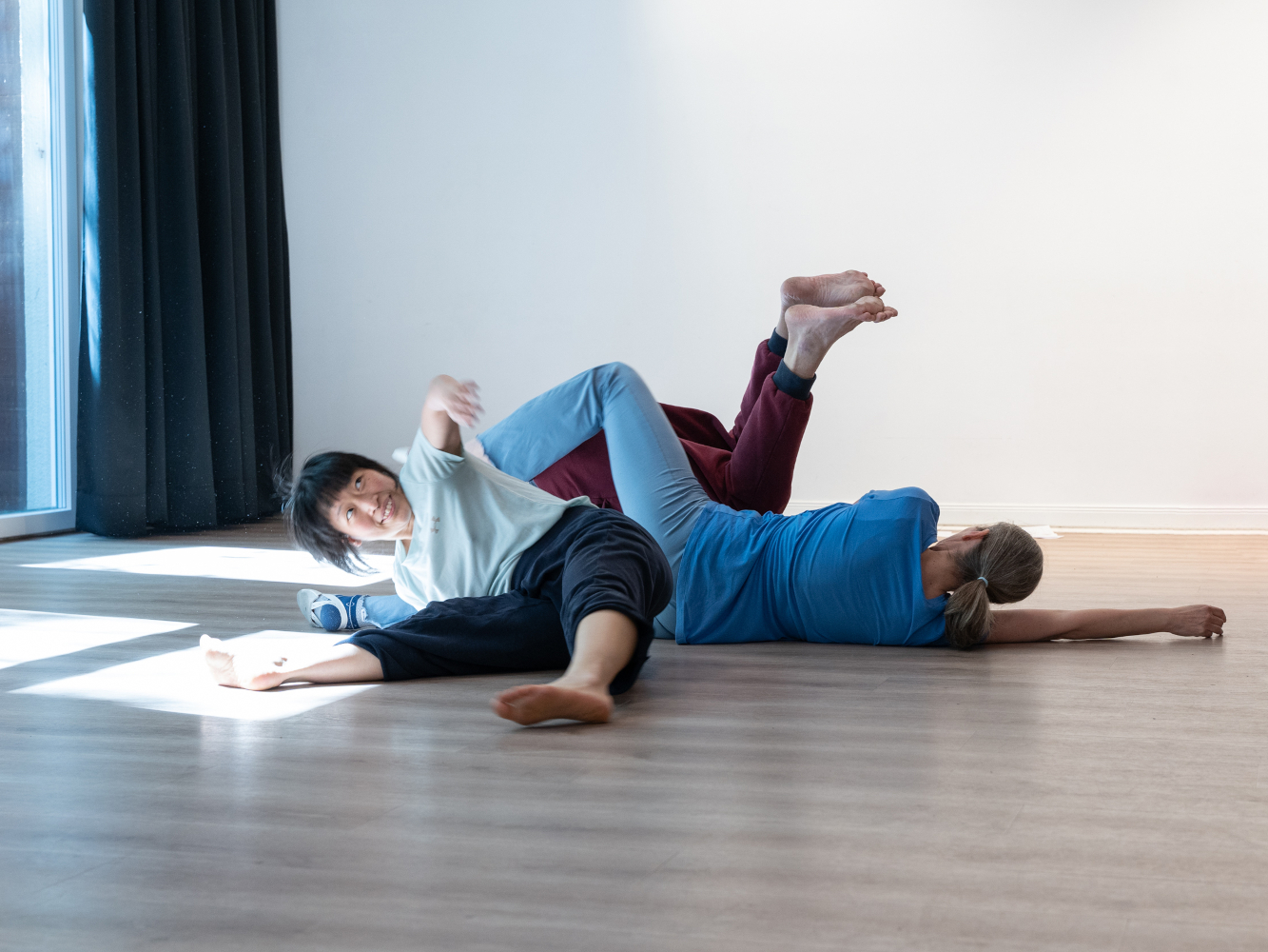 Portrait der Choreografin und Performerin Kai Er Eng. Man sieht Kai bei einer Kontaktimprovisation mit zwei anderen Menschen, sie ist gerade dabei, sich vom Boden wieder aufzurichten. Ihr Blick und ein Arm gehen nach oben zur Decke, sie lächelt. 