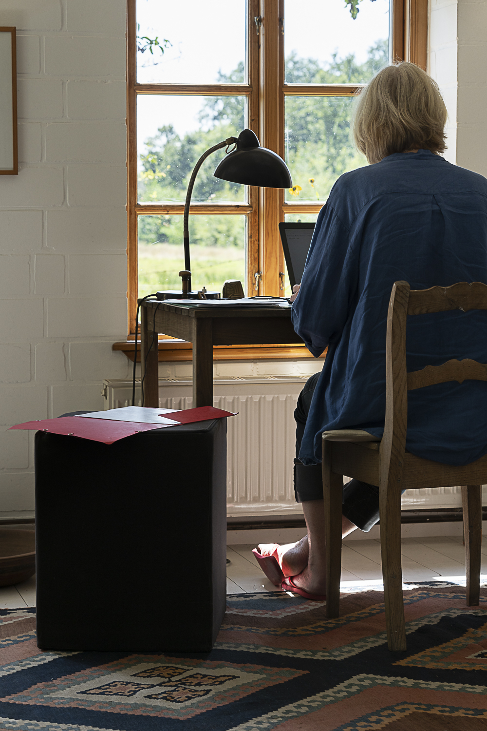 Die Trauerrednerin Stefanie Viereck sitzt an ihrem Schreibtisch vor dem Computer. Man sieht sie von hinten. Sie trägt ein blaues Hemd und hat rote Flipflops an. Durch das Fenster erkennt man ihren Garten.