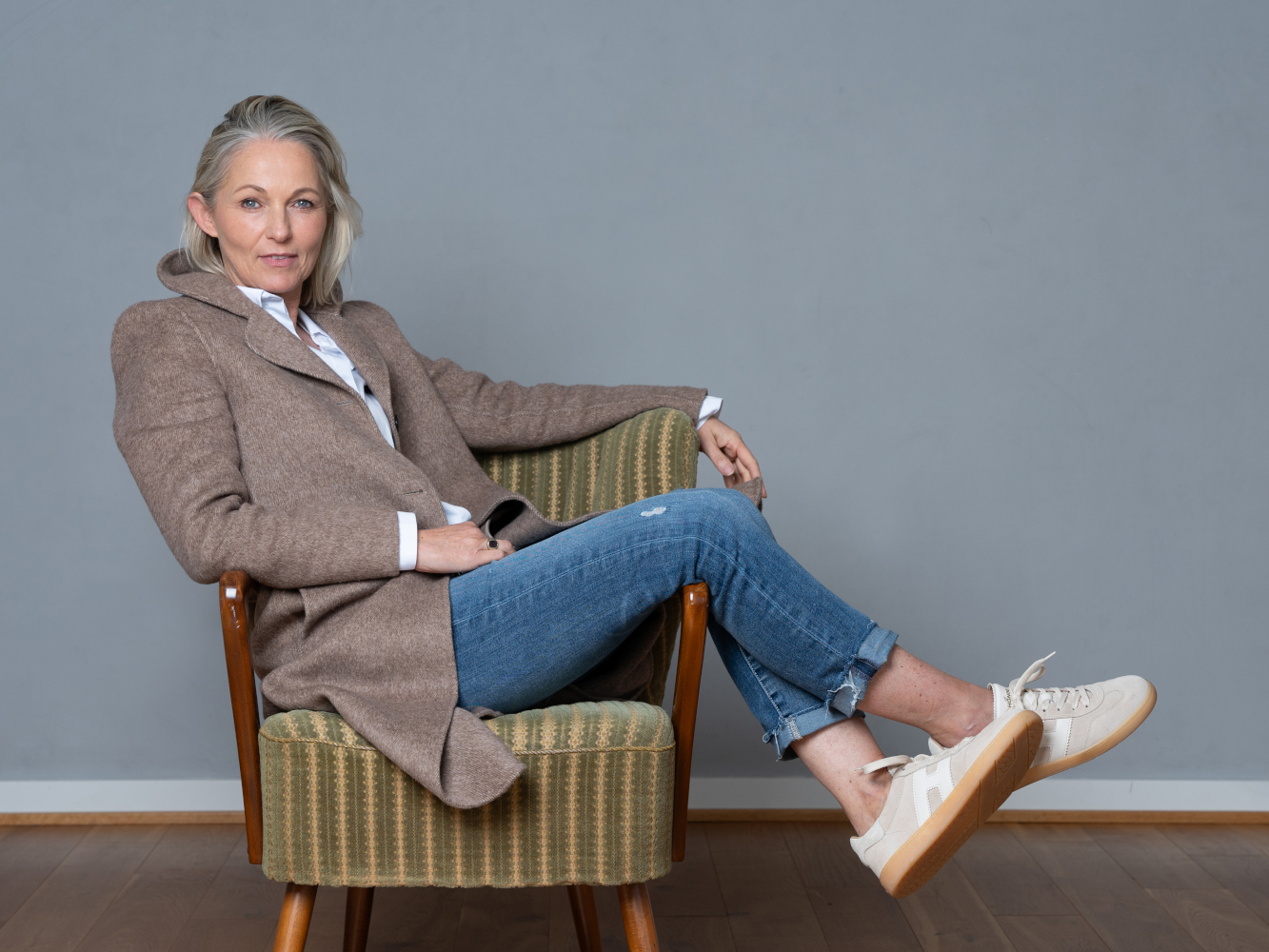 Ganzkörperportrait des Best-Ager-Models Ute Beyer. Sie sitzt quer in einem 50er Jahre Sessel, ihre Beine hängen überkreuzt über der Lehne. Sie hat kinnlange graue Haare, trägt Jeans, einen hellbraunen Mantel und Sneakers. Sie schaut cool und lässig. 