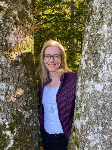 Mental Coach Karin Vogt blick fröhlich zwischen zwei Baumstämmen hervor