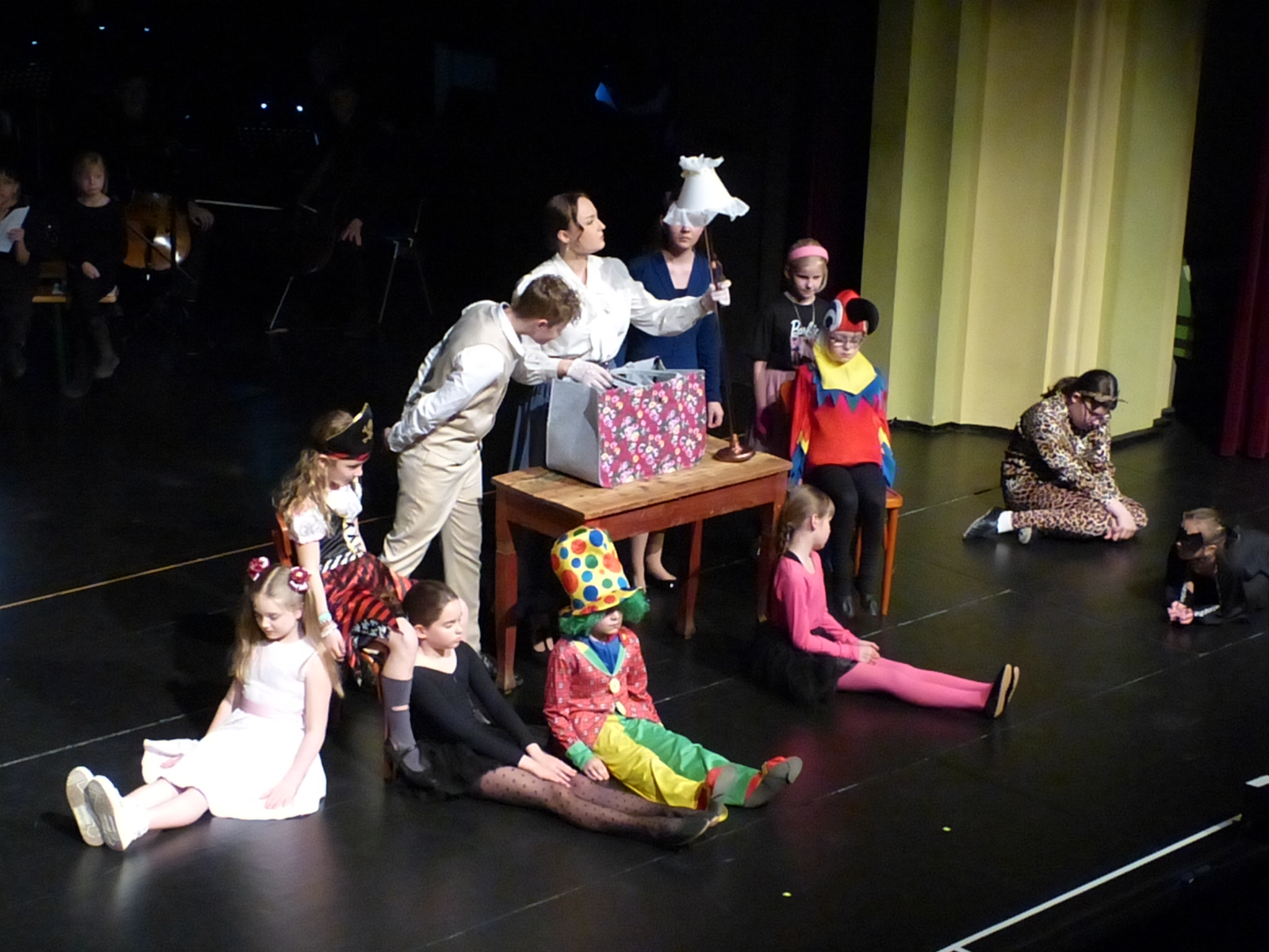 Mary Poppins (Nele Alberti) zieht vor den staunenden Augen von Michael (Bjarne Rose) und Jane (Mara Osten) eine Stehlampe aus ihrer Tasche. Das "Spielzeug", dargestellt von der Nachwuchsklasse des Musiktheaters, "liegt" am Boden.