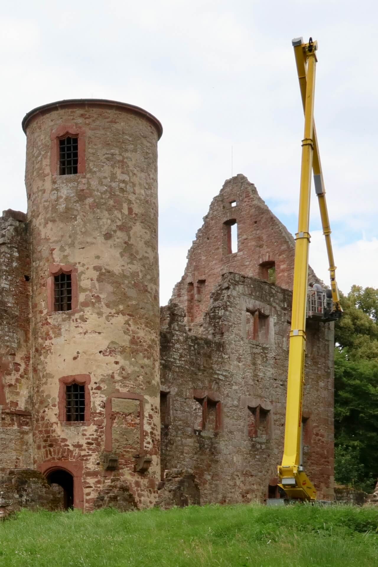 Blick auf die Klosterruine Schönrain im Spessart mit einem gelben Hubsteiger, der an der Ruinenwand steht. Eine Person arbeitet im Korb an der Fassade. Das historische Gemäuer besteht aus rötlichem Sandstein und ist von Grünflächen und Bäumen umgeben.