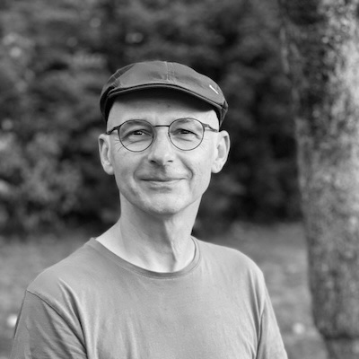 Schwarz-Weiß-Porträt von Christian Söder mit Schiebermütze und runder Brille, der freundlich lächelt. Der Hintergrund ist unscharf. Eine natürliche Umgebung mit Bäumen oder Sträuchern.