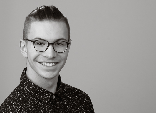 Bewerbungsfoto, ein junger Mann in dunklem Hemd mit dunklen Haaren und Brille posiert für ein Bewerbungsfoto im Querformat