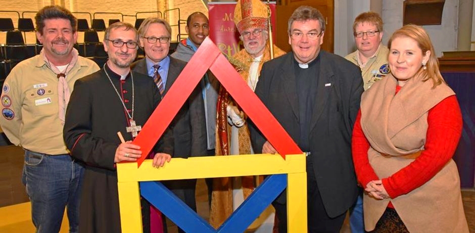 Die im Untertitel erwähnten Leute stehen nebeneinander und schauen in die Kamera. Erzbischof Heße und Msgr. Austen halten ein aus bunten Holzteilen zusammengebautes "Haus des Nikolaus" fest.