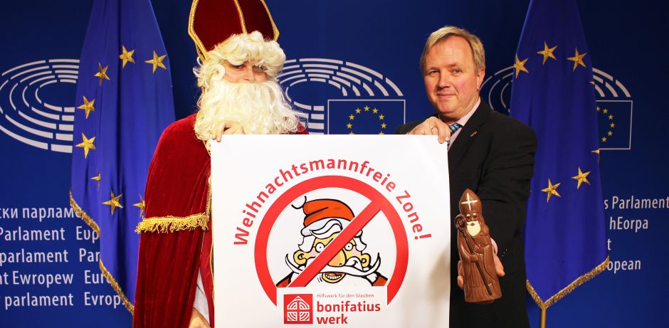 Links steht der heilige Nikolaus und rechts im Bild der Europaabgeordneter Arne Gericke. Zusammen halten sie das Logo der Weihnachtsmannfreien Zone in der Hand. Der Abgeordnete zeigt zusätzlich noch einen Schokonikolaus in die Kamera.