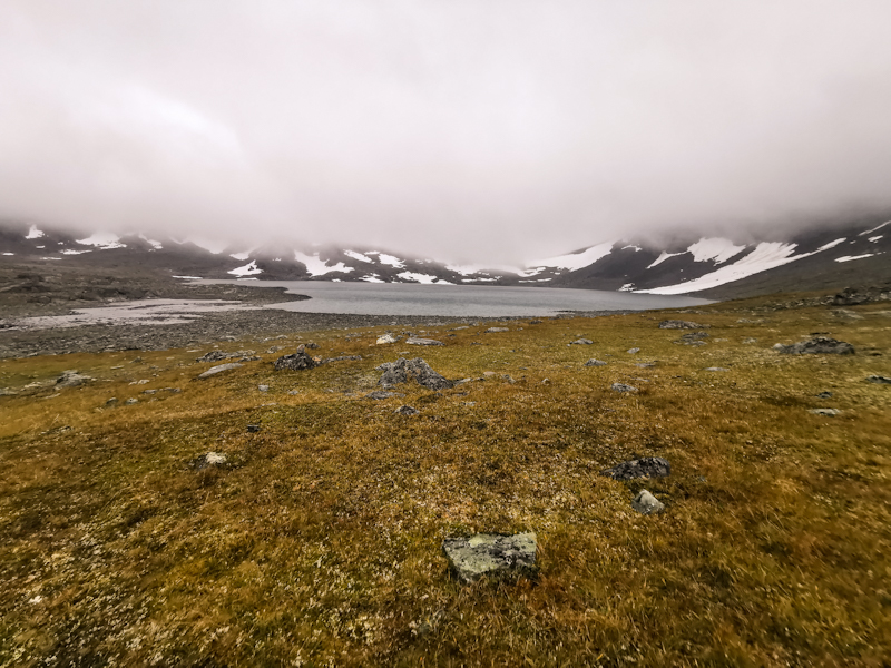 The trail hugs the left side of the lake before climbing through snow fields into the clouds 