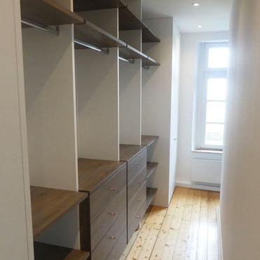 hinter einer Trennwand im Raum ist der Schrank begehbar, die Schränke im Sichtbereich haben Türen, das Mittelteil ist offen gestaltet