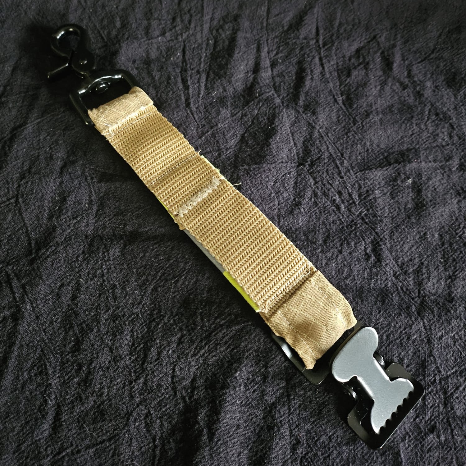 Rückansicht des Handschuhhalters aus beigem Feuerwehr-Gurtband mit stabilem, schwarzem Karabiner und geschlossener Metallschnalle auf Tuch.