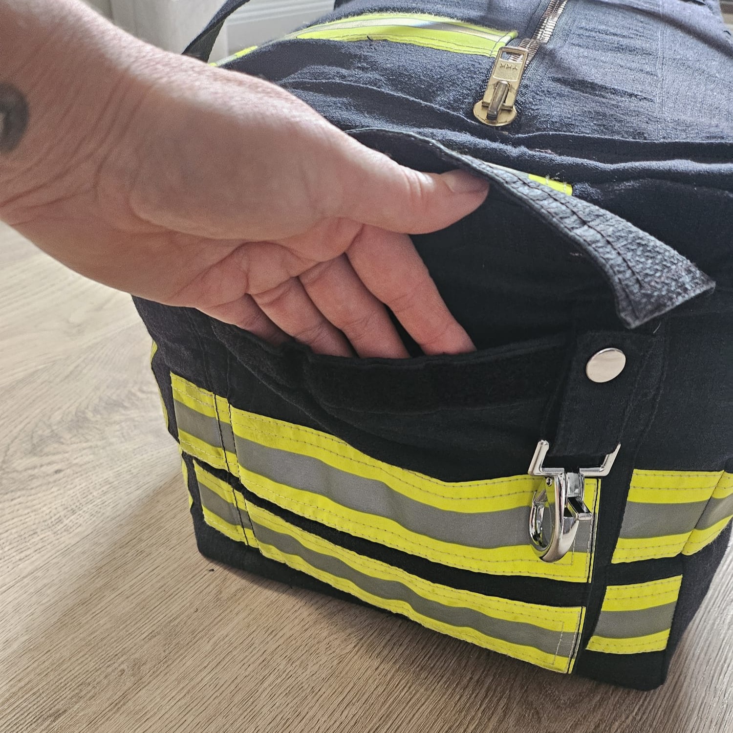 Eine Hand öffnet das Klettfach einer dunkelblauen Feuerwehr-Tasche mit Karabinerverschluss und Reflexstreifen