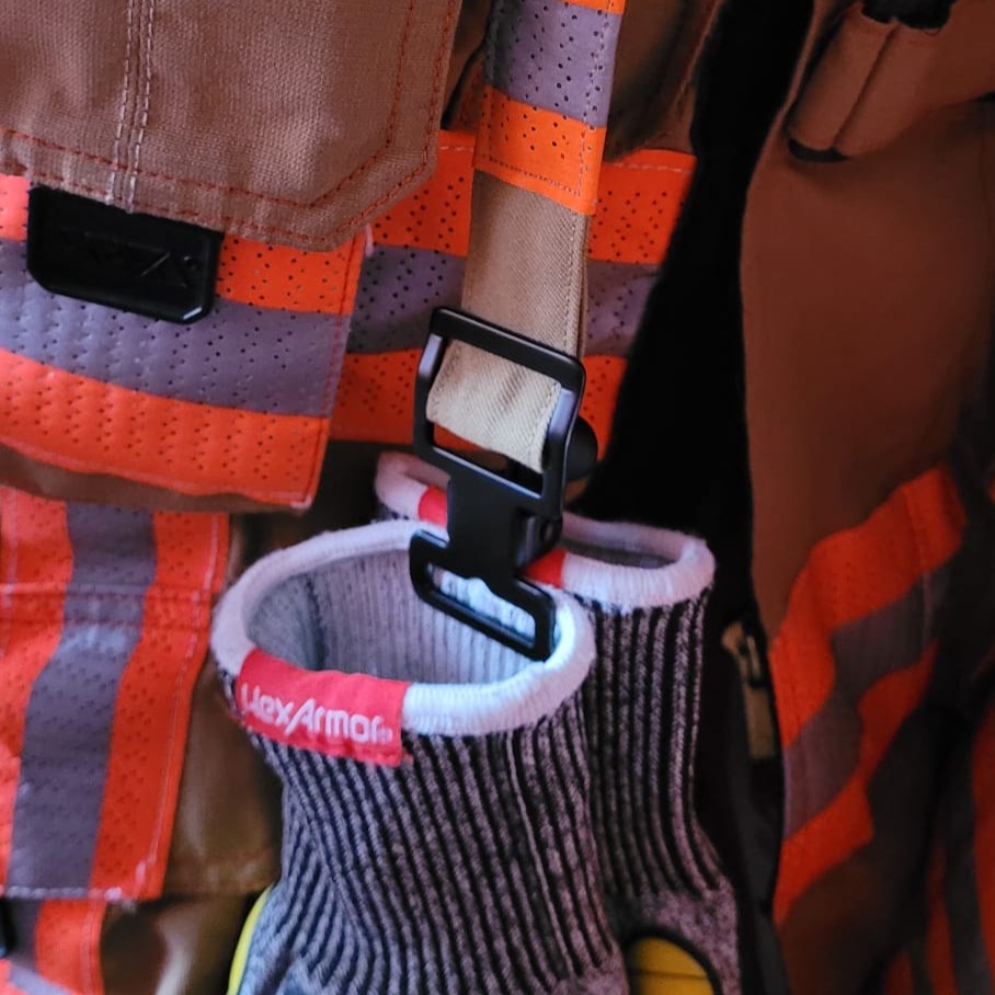 Feuerwehr-Einsatzjacke mit reflektierenden Streifen und eingehängten HexArmor Schutzhandschuhen an einer schwarzen Halterung aus Metall.