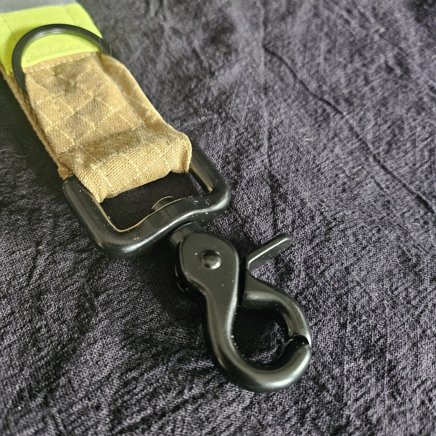 Nahaufnahme eines schwarzen Scherenkarabiner aus Metall, befestigt an einer robusten Stoffschlaufe aus beigem Feuerwehr-Upcycling-Material.