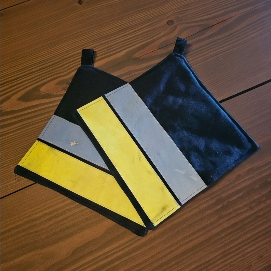 Zwei Feuerwehr Topflappen dunkelblau mit gelben und silbernen Reflexstreifen auf einem Holztisch