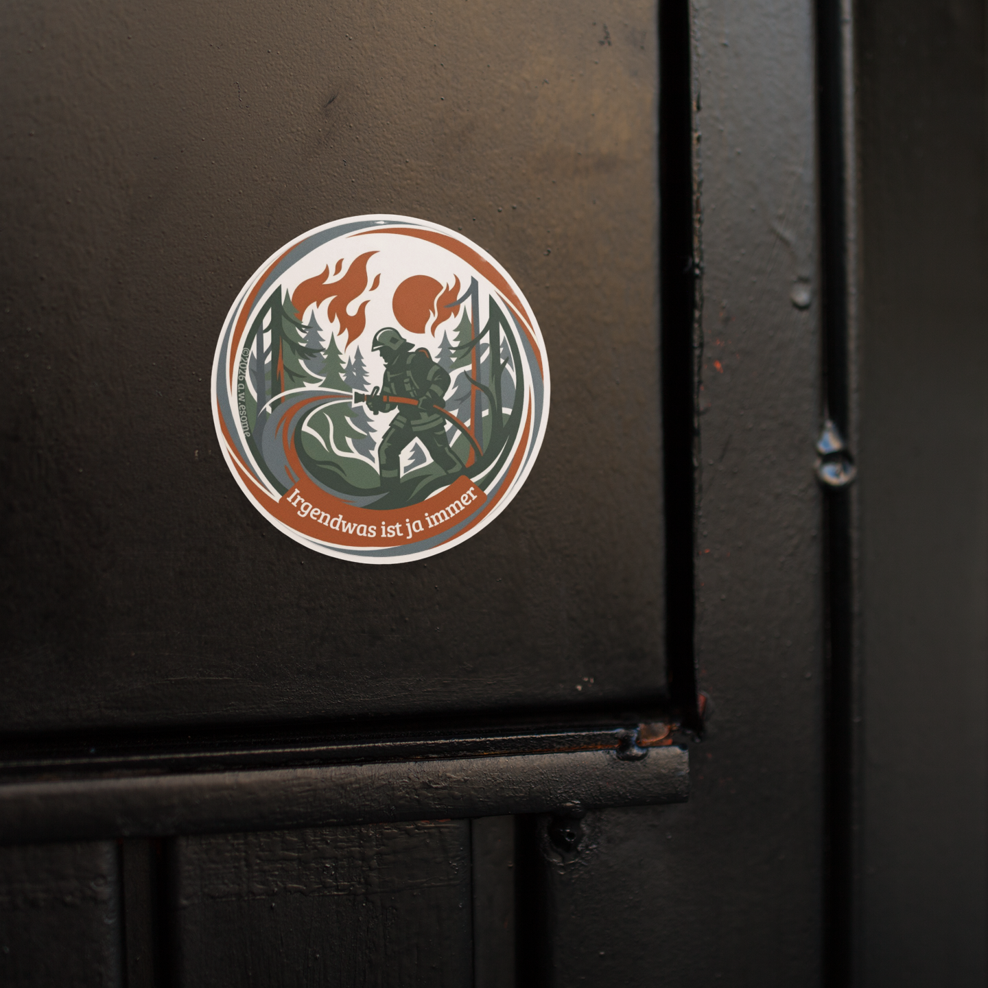 Runder Feuerwehr-Sticker mit Waldbrand-Motiv, aufgeklebt auf einer mattschwarzen Metalloberfläche als praktisches Anwendungsbeispiel.