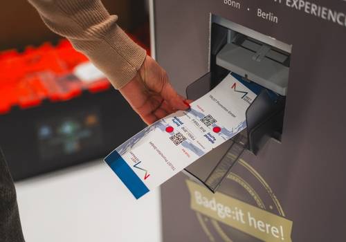 Eine Hand nimmt ein ausgedrucktes Namensschild aus einem Ausgabeschacht. Der Ausgabeschacht gehört zu einem Drucker, der in einem SelfPrint-Kiosk verbaut ist.