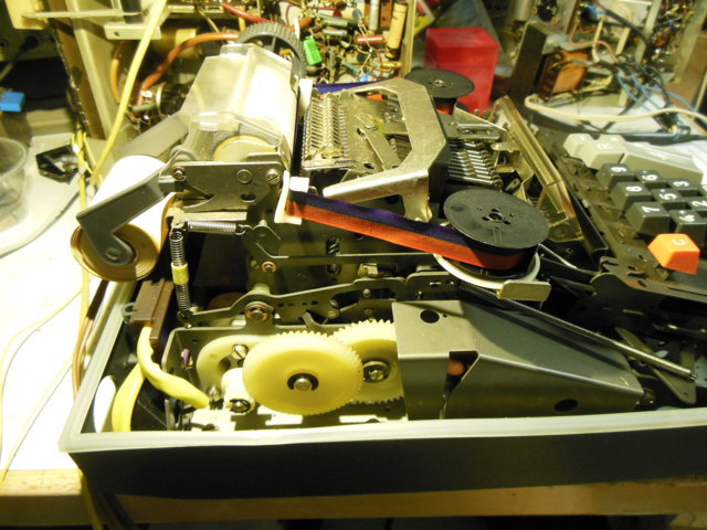 Linke Seite: Getriebe mit Plastikzahnrädern, oben das Druckwerk mit dem Farbband, darunter lang, schräg und silbern die Rückholfeder für den Schlitten.