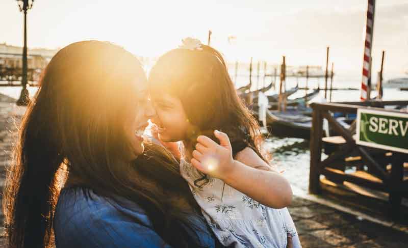 Mom lifting her young child in the air with joyful expressions against a backdrop of traditional Venetian buildings and blue skies.