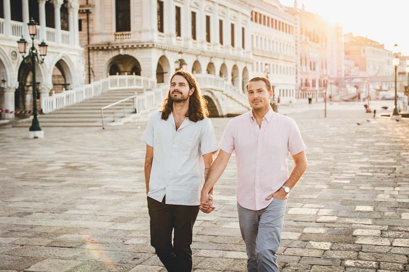 Gay couple holds hands while walking along a Venice canal at sunrise in golden light.