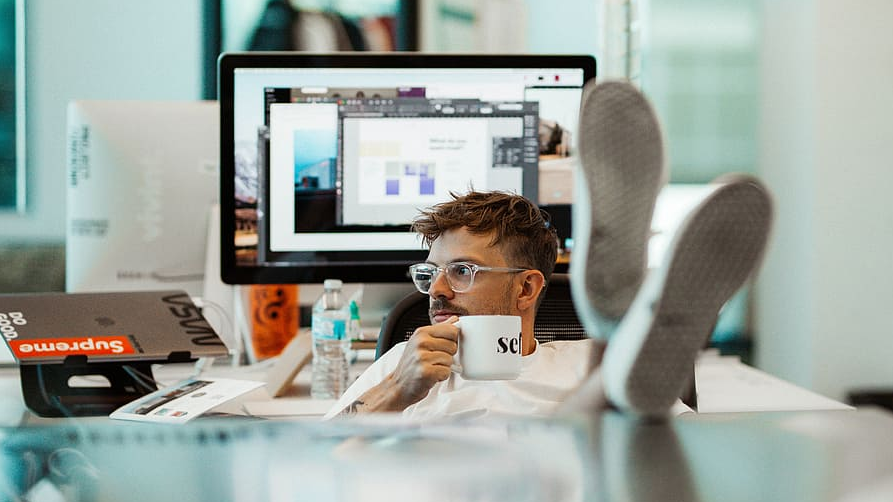 Junger Mann sitzt im Büro mit vollen Schreibtisch, , in der Hand eine Tasse, die Füße auf dem Tisch und sieht aus dem Fenster