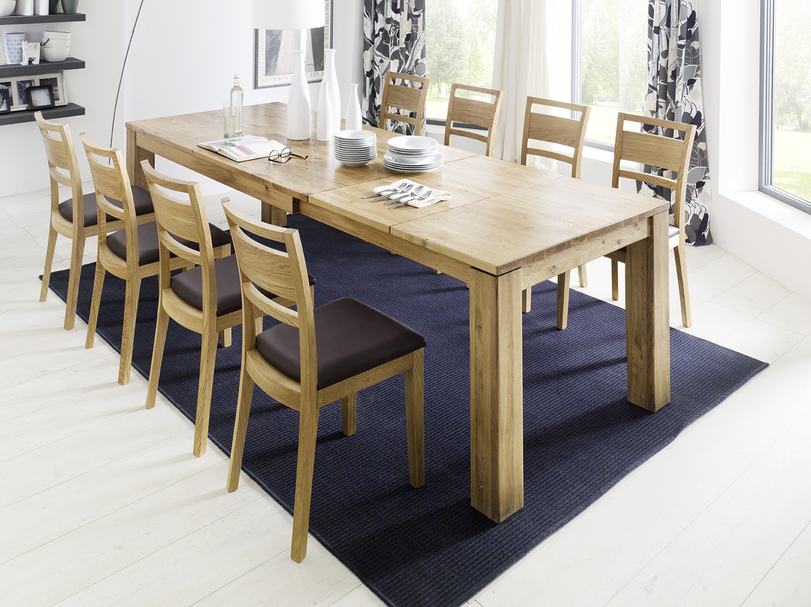 Großer Holztisch mit Ausziehplatte und acht gepolsterten Holzstühlen auf einem dunklen Teppich.