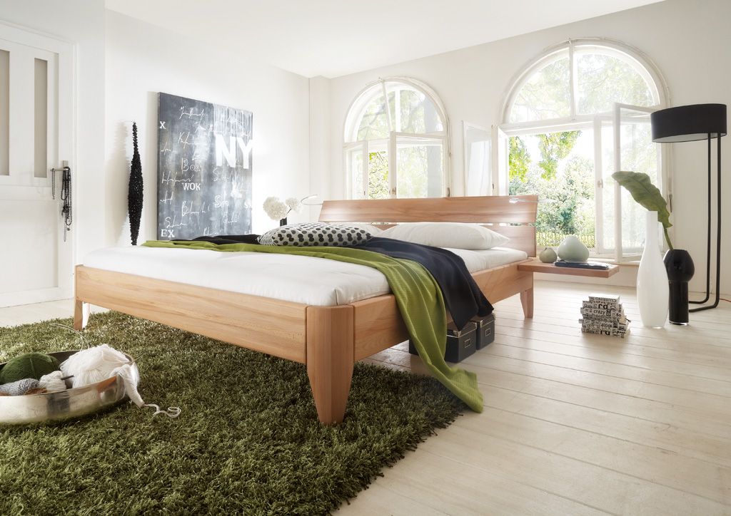 Helles Schlafzimmer mit mittig platziertem Holzbett auf grünem Teppich, gewölbten Fenstern und Kunst.