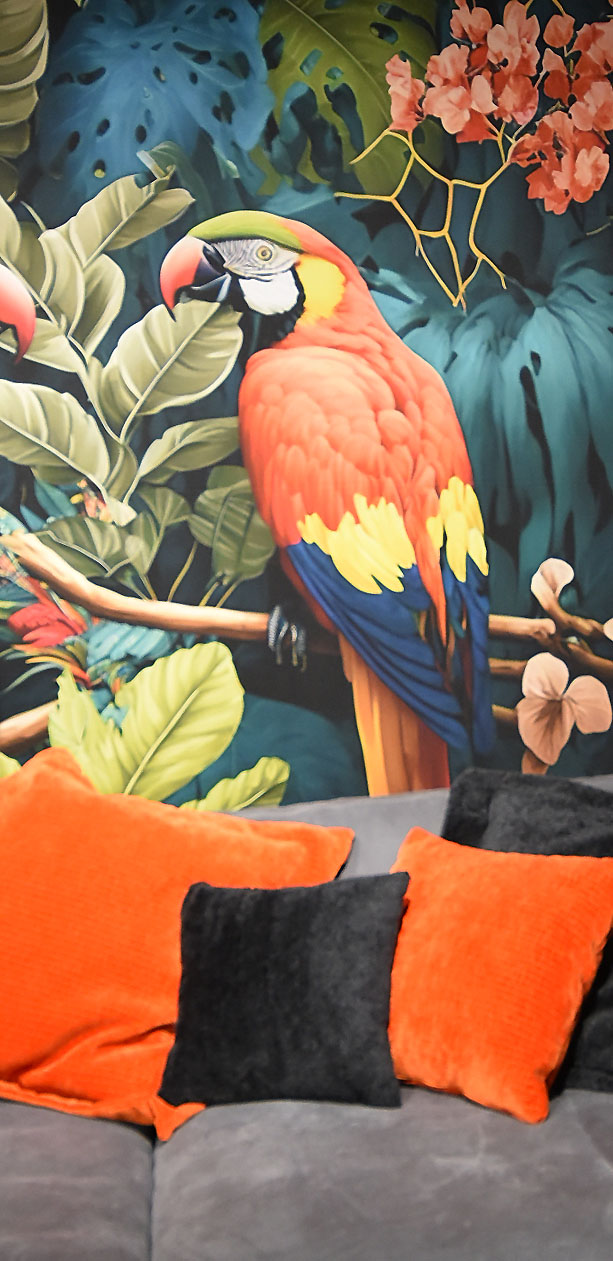 Tropische Tapete mit Papagei und vielen Blättern über einem grauen Sofa mit orange-schwarzen Kissen.