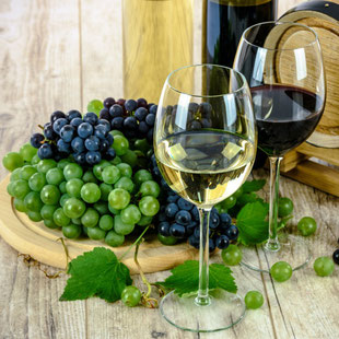 Zwei Weingläser, eines mit Weisswein und eines mit Rotwein, die neben einem Holzbrett voller grüner und blauen Trauben stehen.